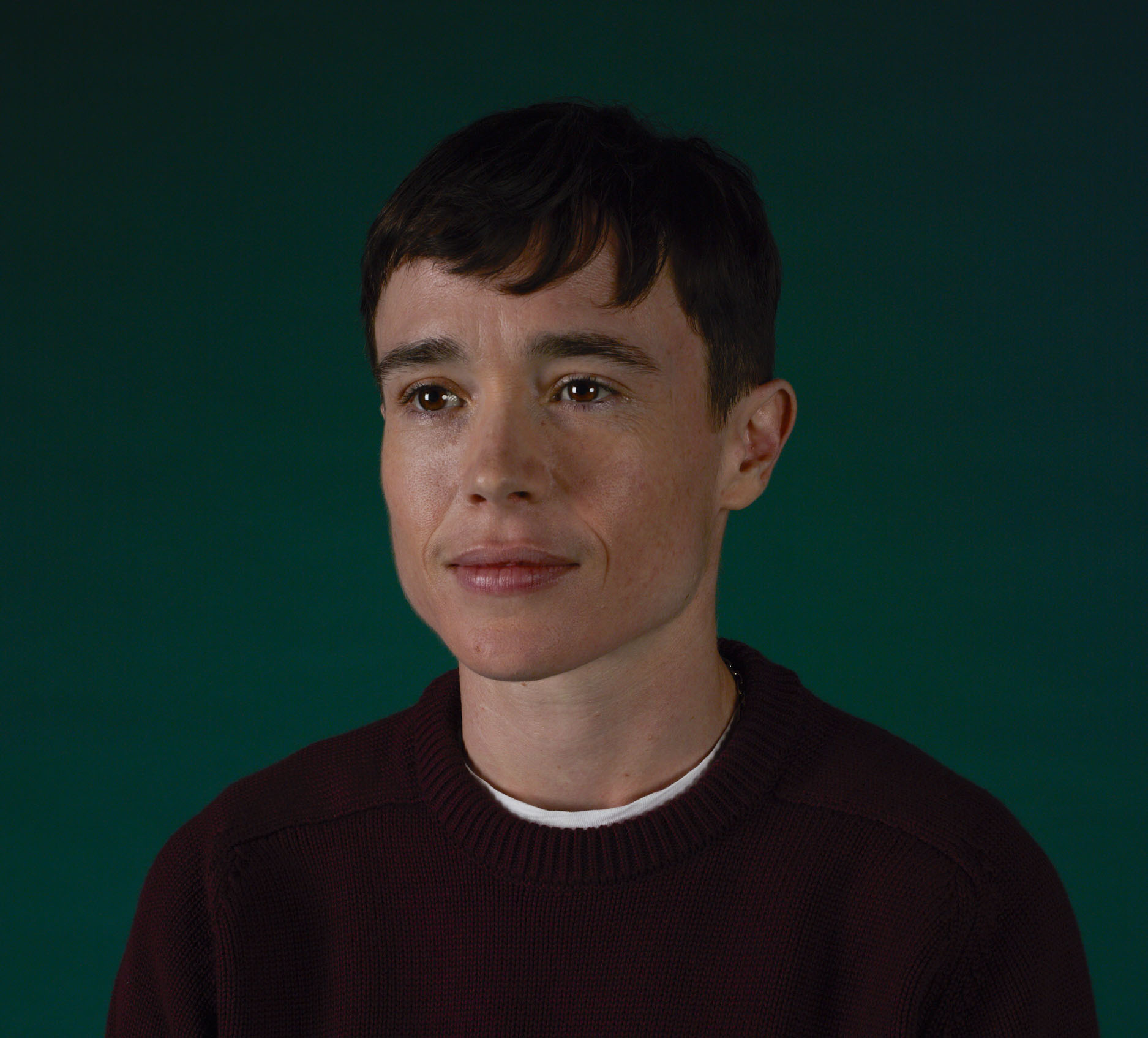 Elliot Page in a black weather against a gree background.