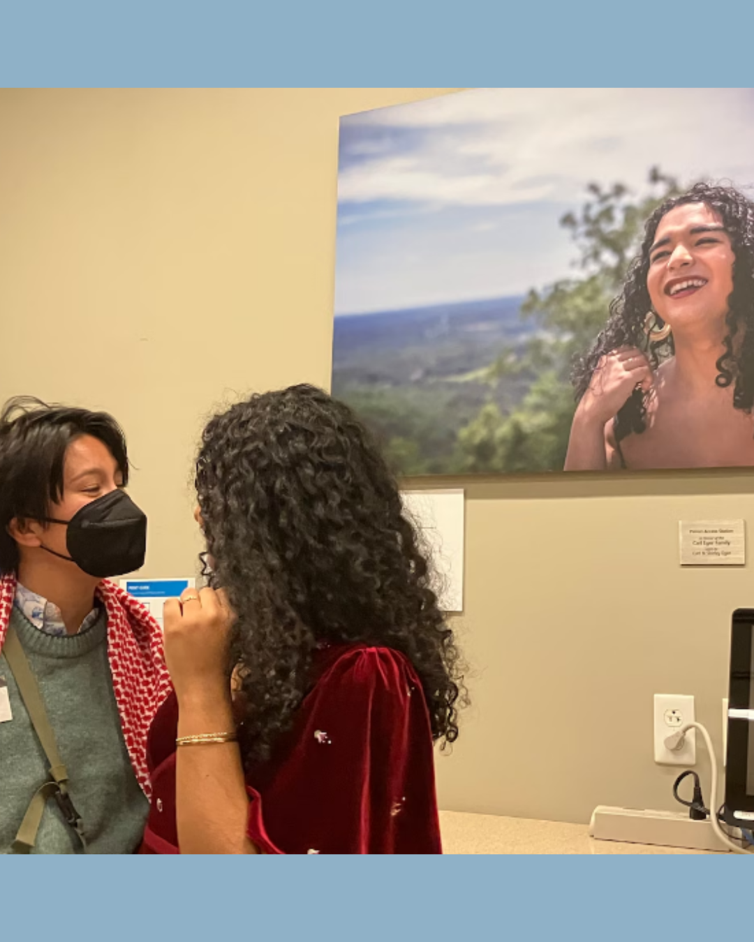 Two people, one with long black curly hair and the other wearing a black mask standing in front of a large photo of a person smiling outside near trees with long black curly hair.