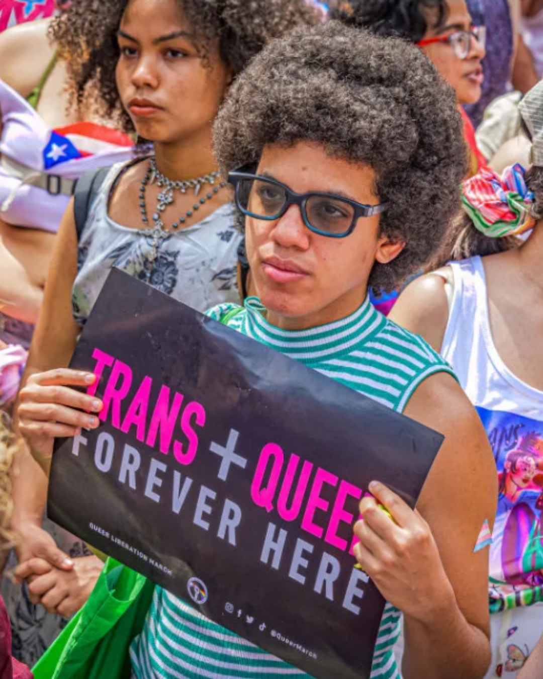 People in colorful clothes standing at a protest, one with a sign that says TRANS + QUEER forever here