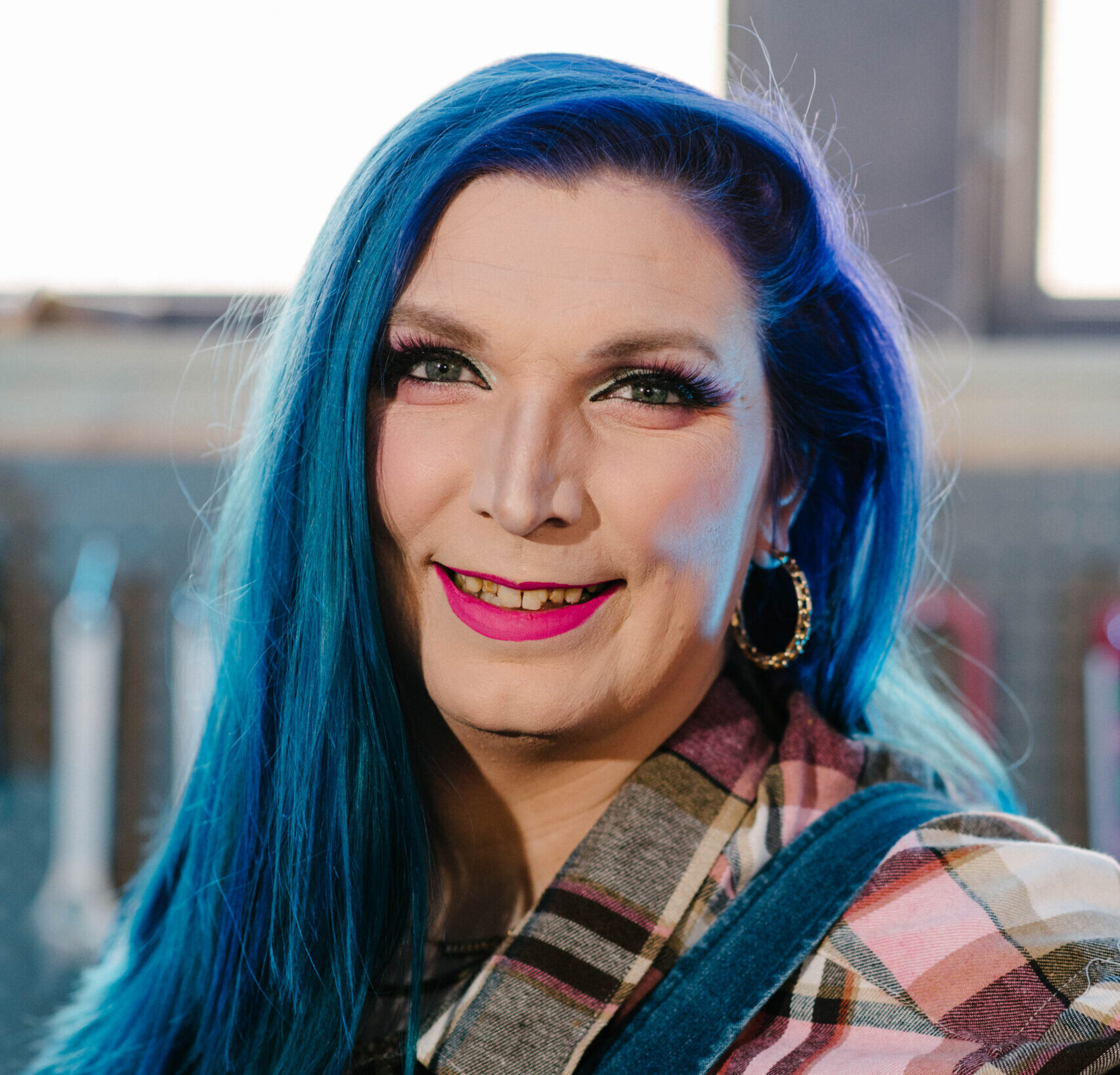 Headshot of Mercury Stardust smiling. She has long blue hair and is wearing denim overalls and a flannel shirt.
